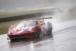 Red race car driving on a wet track with water spray during rainy conditions.