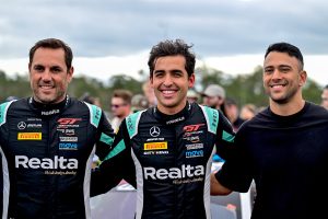 Two race car drivers in racing suits and another man smiling together at a motorsport event.